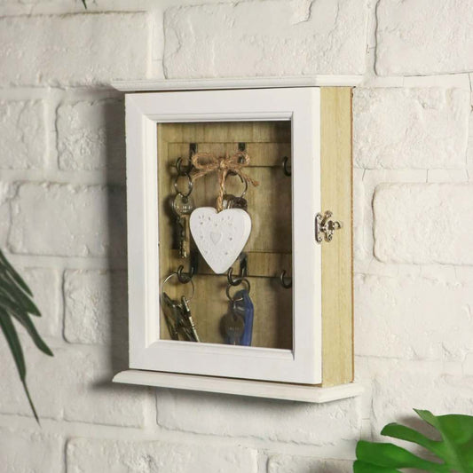 A wall-mounted wooden key cabinet with a white frame and glass door, shown holding several keys and a white heart-shaped ornament against a white brick wall.