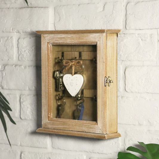 A rustic wooden wall-mounted key cabinet with a glass door, hanging keys and hooks, and a white heart-shaped ornament, shown against a white brick wall.