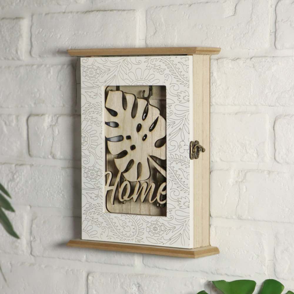 A wooden wall-mounted cabinet with a white patterned door and a central cutout showing a monstera leaf design and the word 'Home' in wood, mounted on a white brick wall.